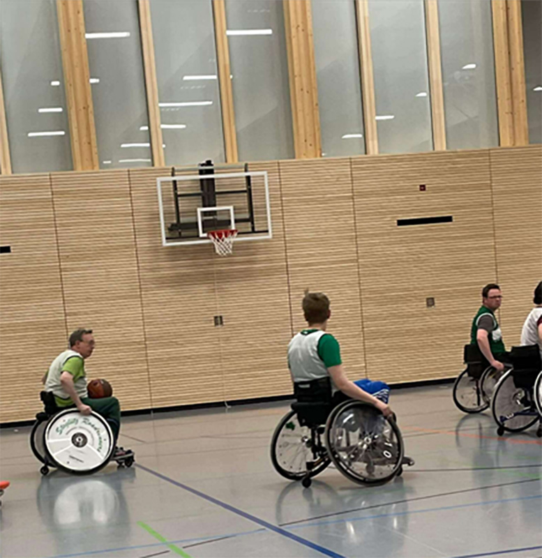 Vier Personen in Rollstühlen spielen in einer Sporthalle Basketball.