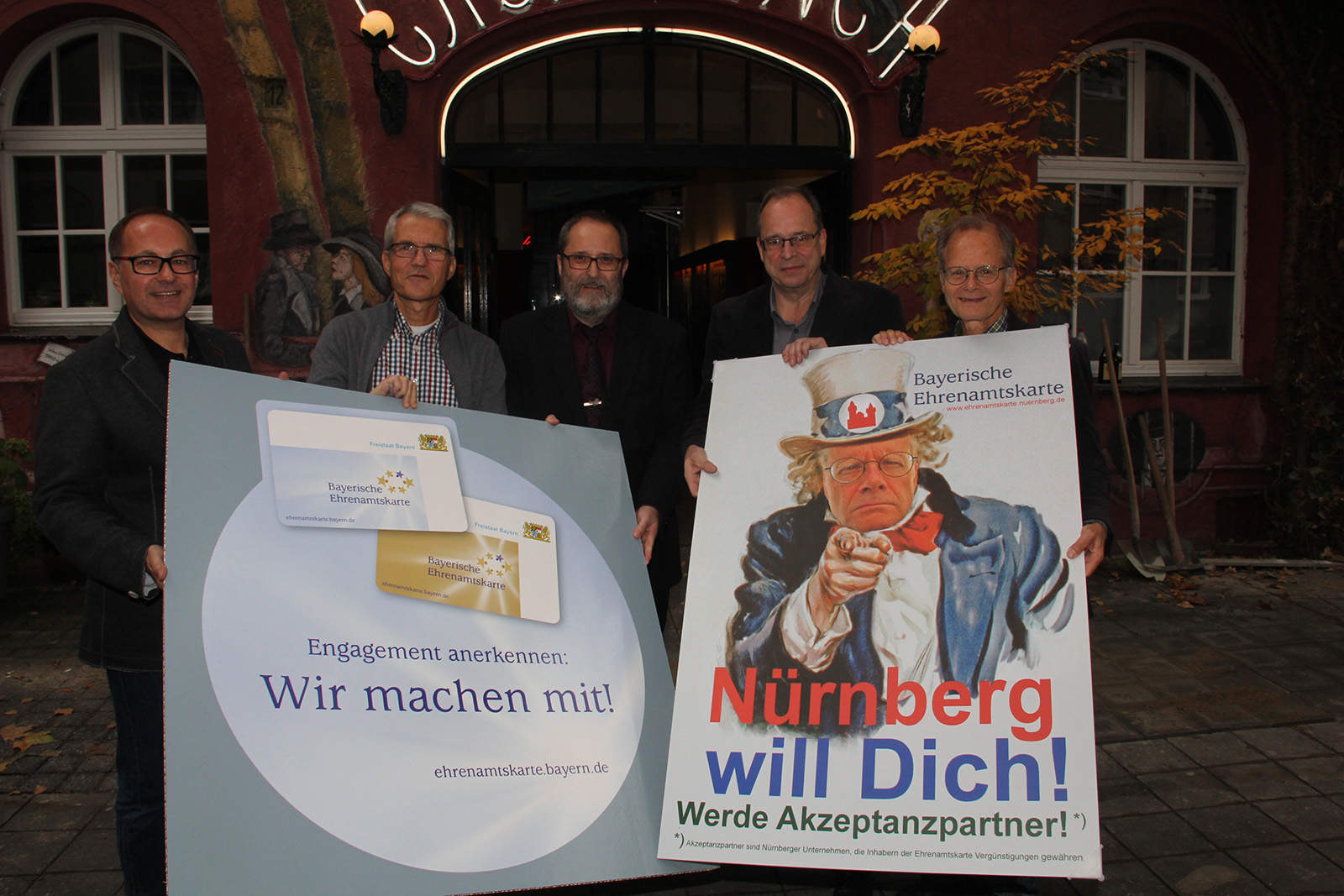 Fünf Männer halten große Plakate über bayerisches Ehrenamt vor einem Restaurant.