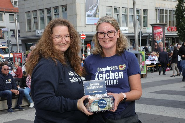 Zwei Frauen stehen lächelnd auf einer belebten Straße und halten gemeinsam eine Auszeichnung.