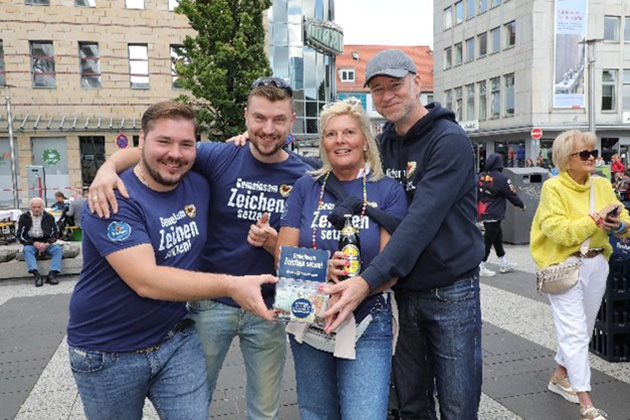 Vier Personen posieren lächelnd und halten gemeinsam ein Buch auf einer belebten Straße.