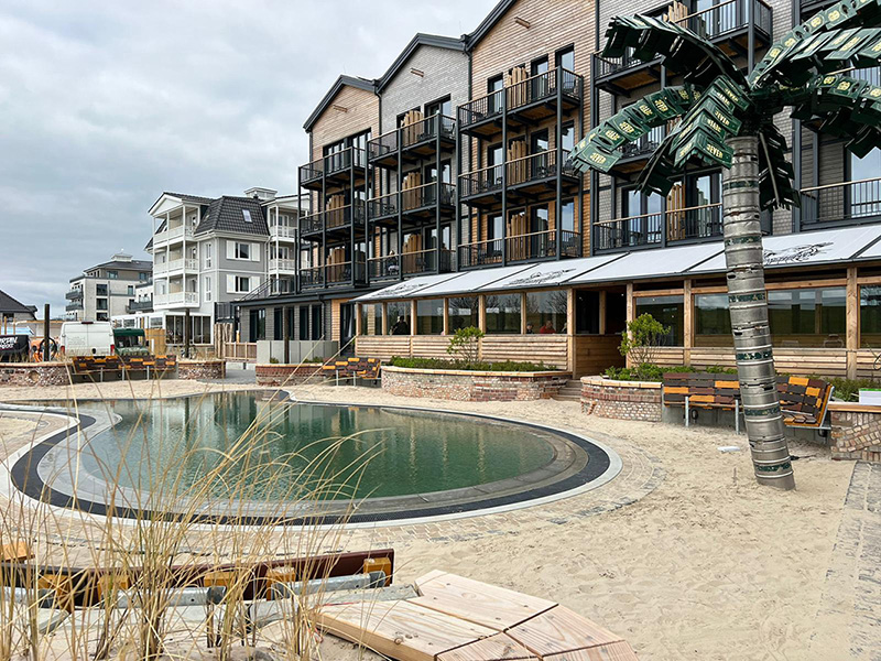 Ein modernes Strandresort mit mehrstöckigen Holzgebäuden und einem zentralen Teich. Eine künstliche Palme steht neben einer Terrasse.