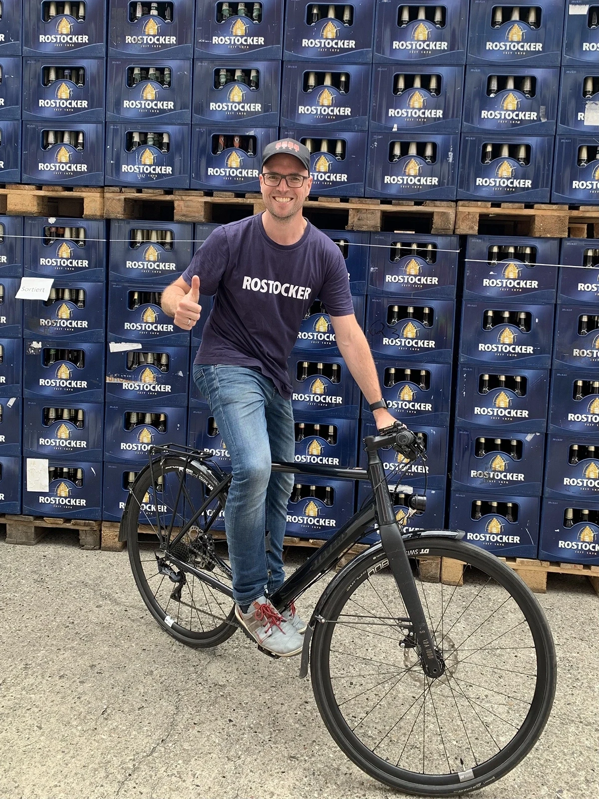 Ein lächelnder Mann steht vor blauen Kisten und lehnt an einem Fahrrad. Er trägt eine Brille und zeigt den Daumen hoch.