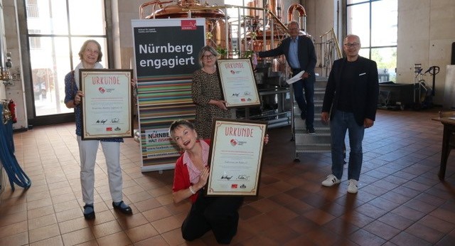 Fünf Personen stehen in einer Brauerei und halten freudig Urkunden.