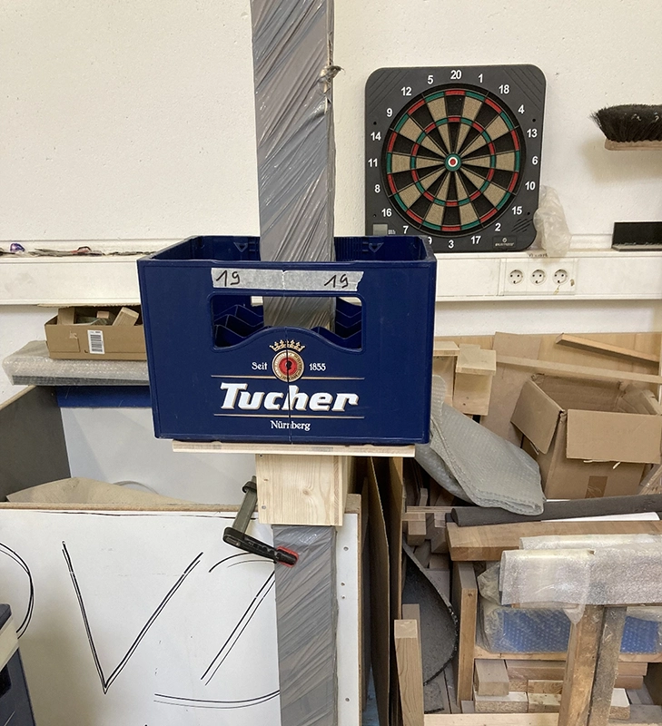 Eine Getränkekiste steht im Vordergrund vor einer Werkstattwand mit Dartscheibe.