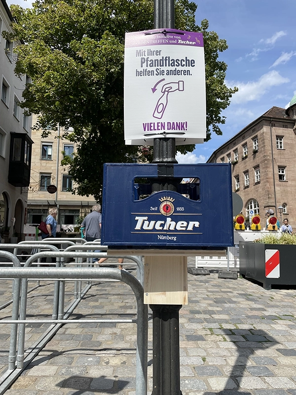Ein Schild fordert dazu auf, Pfandflaschen zu spenden, montiert auf einem Müllbehälter in einer belebten Straße.