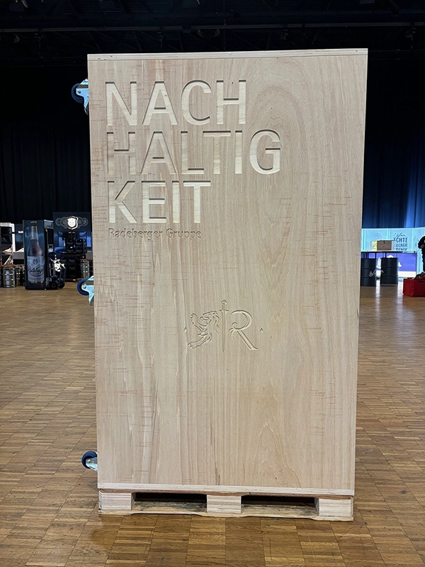 Ein Holzpodium mit dem Wort NACHHALTIGKEIT und einem Firmenlogo darauf steht in einem Saal.
