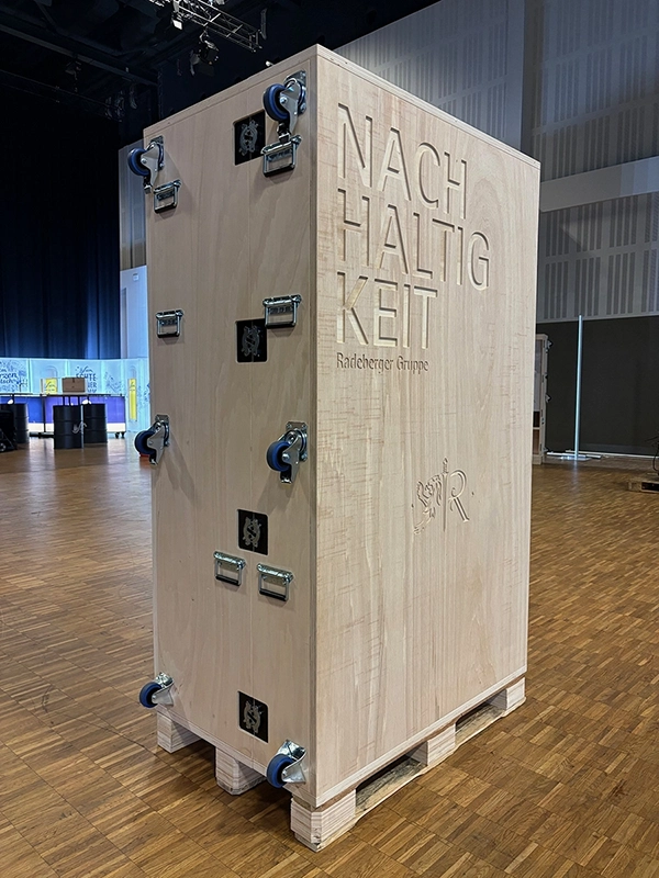 Große, hölzerne Transportkiste mit Rollen steht auf einer Bühne, das Wort Nachhaltigkeit ist eingraviert.