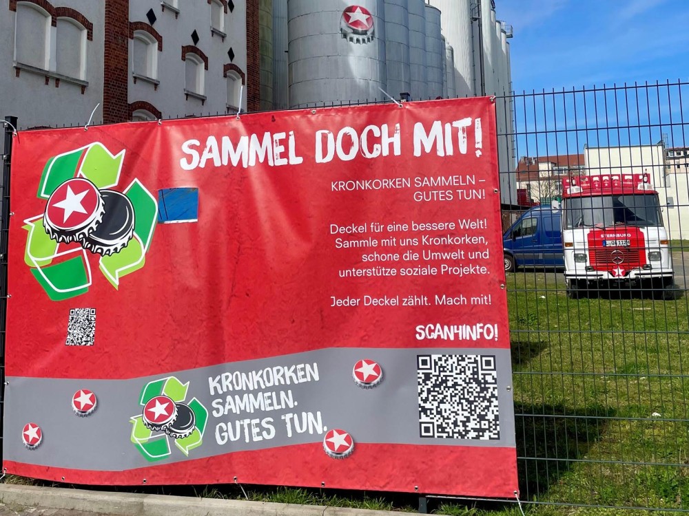 Ein rotes Werbebanner wirbt für das Sammeln von Kronkorken, um die Umwelt zu schützen und soziale Projekte zu unterstützen.