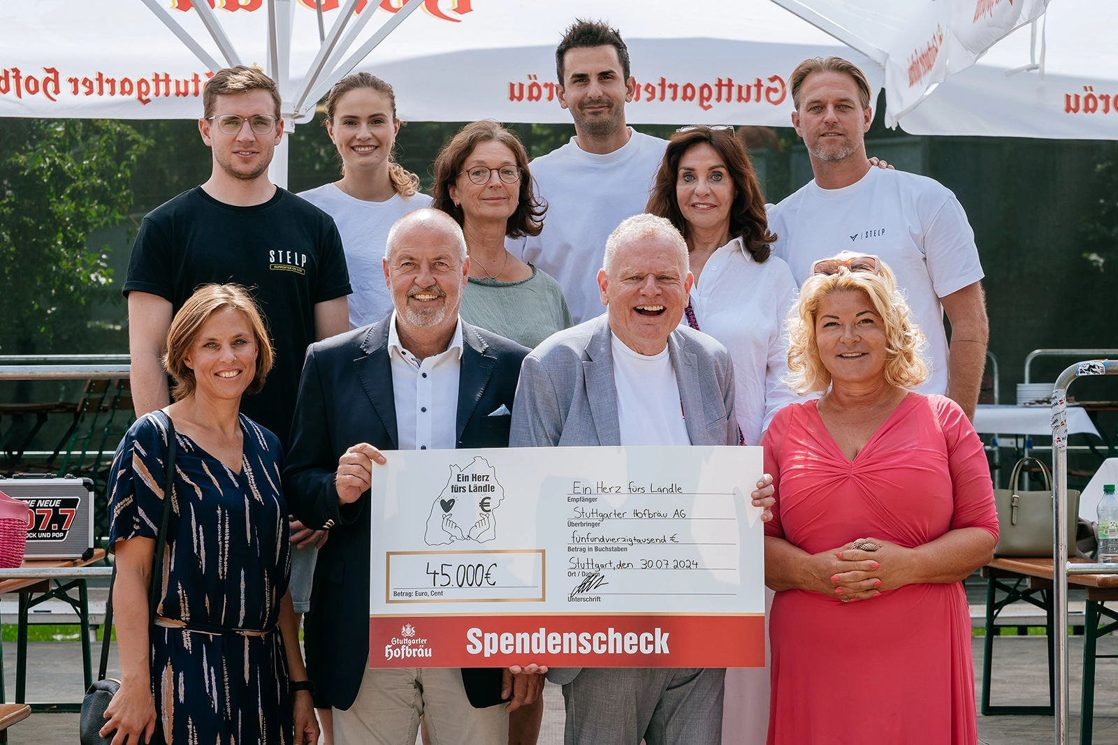 Gruppe von zehn Personen hält zusammen einen großen Scheck bei einem Event im Freien.