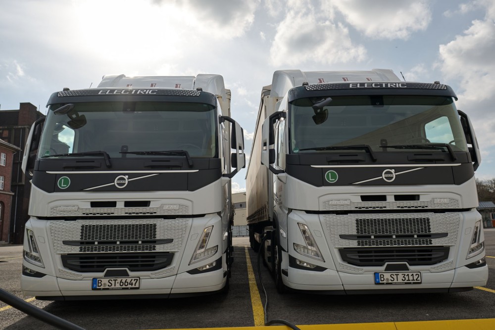 Zwei elektrische Lkw stehen nebeneinander an einer Ladesäule. Sie sind weiß und tragen Kennzeichen aus Berlin.